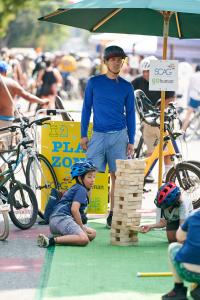 Kids playing giant Jenga outdoors; adult supervising, wearing blue.